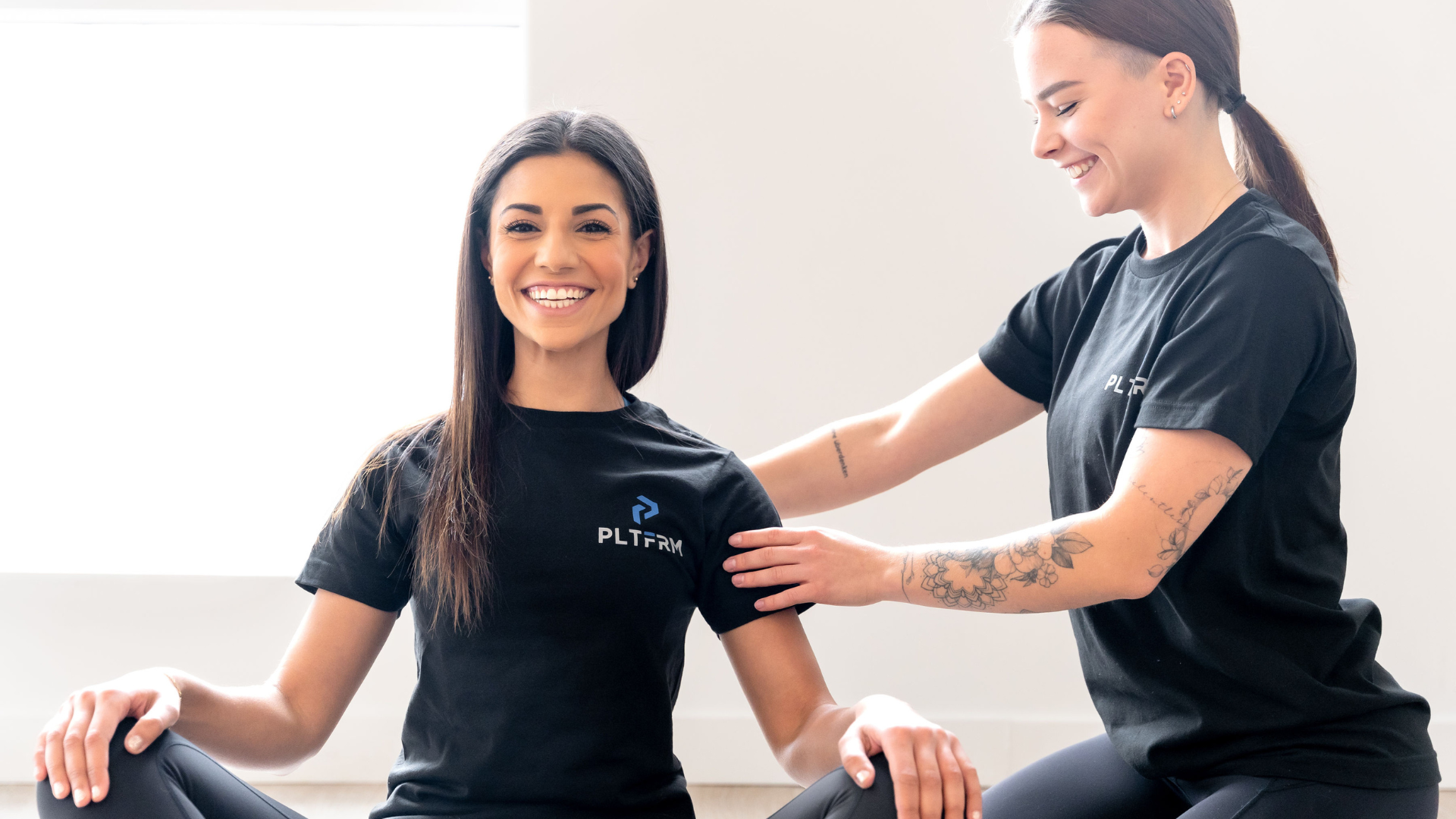 Two personal trainers in PLTFRM t-shirts. One trainer is adjusting the other in a seated yoga pose.